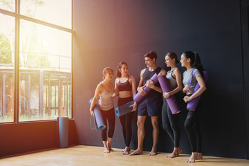 Group of young sporty man and women with yoga mats standing at black wall smiling and talking after...