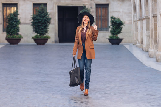 Excited female in stylish casual wear and black hat walking and looking at camera on sidewalk among old buildings