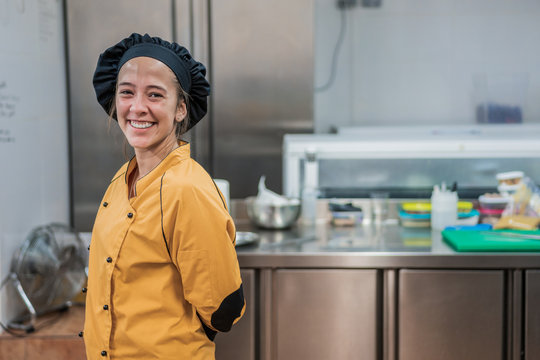 Cheerful young female chef in yellow uniform and black hat looking at camera while  standing in restaurant kitchen