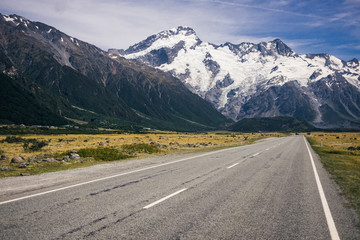 Asphalt road amid tropical green cliffs with cloudy blue sky and mountain Cook with snow on top on the background