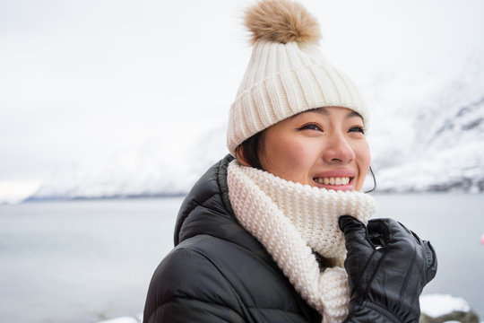 Happy female with amazing eyes in black jacket and white warm hat and scarf looking away and smiling against white blurred background in Norway