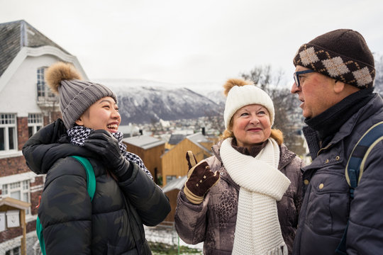 Pleased Asian female on vacation in warm clothing laughing while talking with local citizens at city street at Tromso in Norway