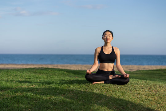 Young slim female in black top and leggings sitting on lotus position on green grass with closed eyes while meditating at the beach