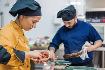 Side view of young female cook in yellow uniform holding pot with sauce while working together with male colleague in restaurant kitchen 