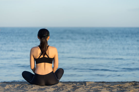 Back view of young fitness female in black sportswear sitting with crossed legs on beach resting after workout