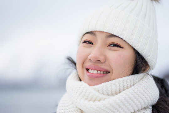 Happy female with amazing eyes in black jacket and white warm hat and scarf looking at camera and smiling against gray blurred background in Norway