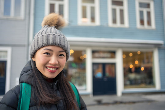 Gorgeous adult Asian female in warm clothes looking at camera and smiling against blurred exterior of blue modern building in downtown