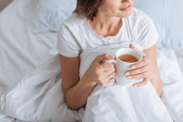 cropped view of dreamy woman holding cup with tea in bed