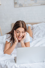 excited young woman using laptop while chilling in bedroom