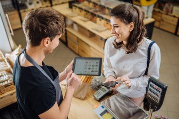 Small business, people, takeaway and service concept. Happy man owner stands behind counter giving paper bag to female customer at zero waste bakery grocery shop.
