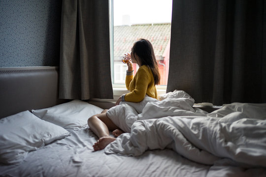 Side view of black haired faceless female in yellow sweater sitting on bed while drinking tea and looking at window on lazy morning at Tromso in Norway