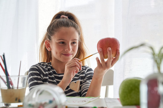 Cheerful girl smiling and coloring fresh apple while sitting at table and painting at home