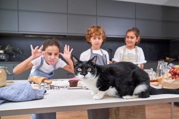 Shocked male and female kids in aprons looking at big cat on table with ingredients at modern kitchen