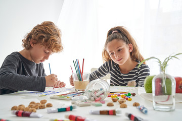 Concentrated girl and boy in casual outfit painting with watercolor while sitting at table at home