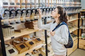 Minimalist vegan style young woman with cotton reusable net mesh bag on background of goods of zero waste shop. Shopping without plastic packaging in plastic free grocery store.