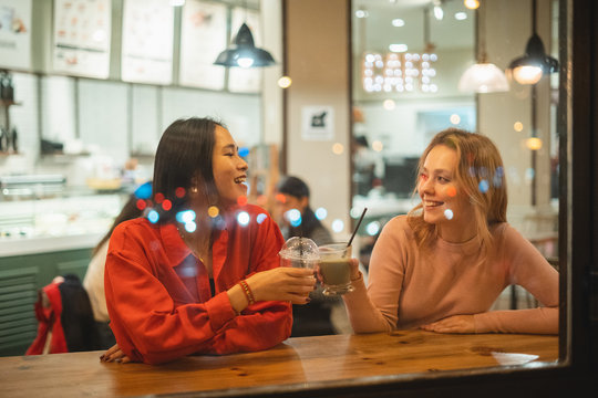 Young multiracial women smiling and speaking with each other while sitting at table in cozy cafe clinking cups and enjoying fruit drinks