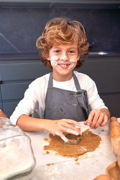 Content boy with face painting dirty flours trying to mold brown paste on table with cooking things at kitchen