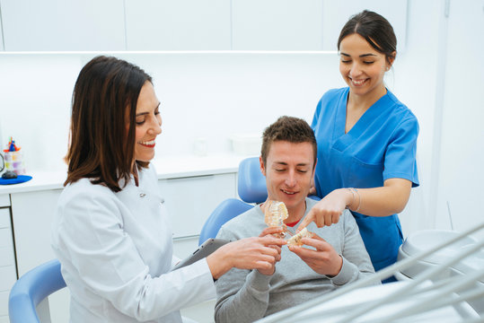 Enthusiastic patient in chair holding prosthesis and dentist with assistant in uniform explaining teeth structure