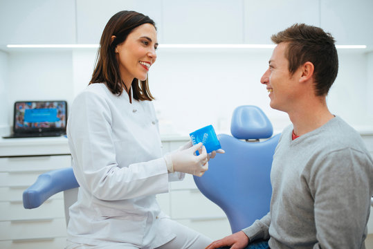 Cheerful qualified dentist and patient discussing process of treatment and doctor showing prosthesis in blue box in cabinet