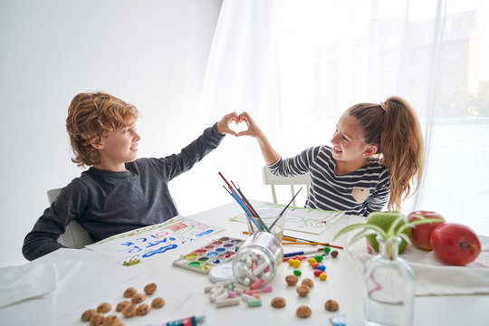 Little girl and boy smiling and showing heart gesture while sitting at table and painting at home together
