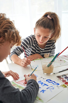 Concentrated girl and boy in casual outfit painting with watercolor while sitting at table at home