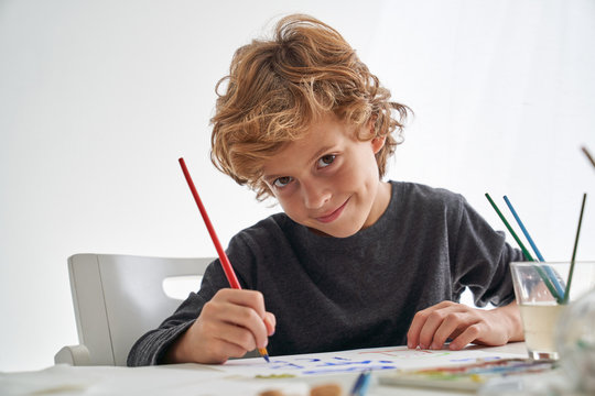 Little boy with curly hair smiling and looking at camera while sitting at table and painting against white wall at home