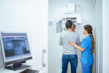 Back view of client making x-ray with help of careful assistant in blue uniform in dental cabinet with monitor and equipment