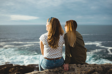 Back view of young sisters in casual clothes dreaming and enjoying sunrise while sitting together on rocky coast with ocean waves breaking on shore and cloudy sky in background