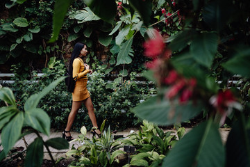 Side view of trendy Asian woman with long dark hair in yellow shirt and short skirt walking in beautiful garden and looking away
