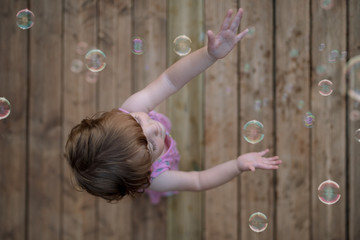 From above adorable child in pink dress laughing and capturing rainbow soap bubbles on wooden ground