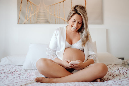 Content blonde pregnant female in white open shirt and bra browsing mobile phone while sitting with crossed legs on big bed in bright room