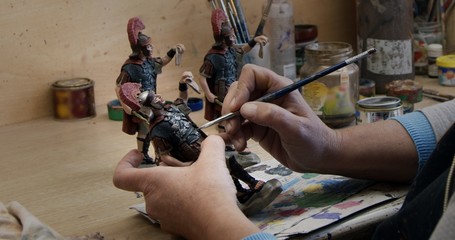 Side view of crop person painting clay figurines of soldiers with brush at table in pottery
