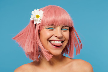 Pleased young beautiful pink haired woman with colored makeup having nice time while posing over blue background, showing happily her tongue and keeping eyes closed