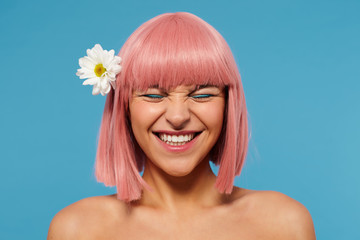 Portrait of young beautiful pink haired woman with bob haircut showing her perfect white teeth while smiling happily with closed eyes, isolated over blue background