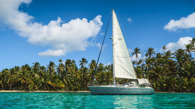 A grey-white Sailing Yacht anchored in Turquoise Water in front of the paradisiacal San Blas Islands in Panama with green Palm Trees and a perfect Blue Sky in the background.