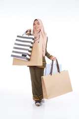 A beautiful Muslim female college student in a Asian traditional dress modern kurung carrying shopping bags isolated on white background. Eidul fitri fashion and festive shopping concept.