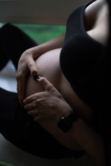 Pregnant woman in black clothes holds hands on belly on a dark background. Pregnancy, maternity, preparation and expectation concept - close up of happy pregnant woman with big belly at window.