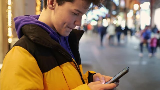 Teenage boy typing text message and communicate in social networks using his smartphone. Smiling boy on a busy city street with citylight in the evening. UHD, 4K