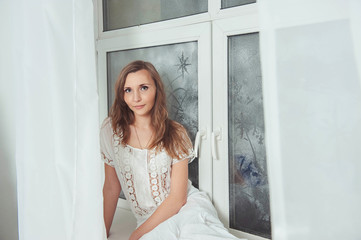 Portrait of charming sensual beautiful and sexy girl in white dress sitting on window. Young beautiful woman waiting for spring in white light room.