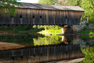 Old New England covered bridge over calm river. Reflection of bridge in water during autumn season