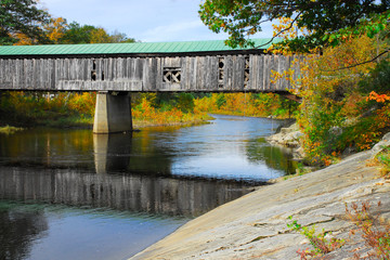 Old New England covered bridge over calm river. Reflection of bridge in water during autumn season