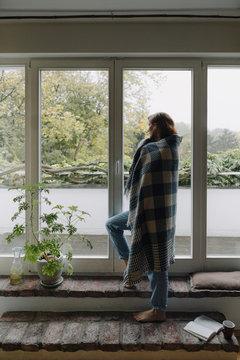 Woman looking out of window, wrapped in blanket