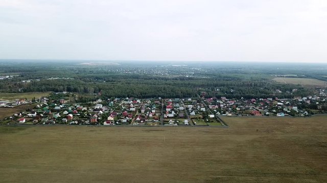 View of the small provincial town located in ecological area near green forest and field on cloudy sky background. Stock footage. Living in countryside, houses surrounded by meadow and trees.