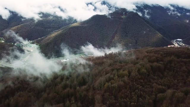 Autumn in foggy mountains covered by trees and ski resort in ecological area. Stock footage. Aerial of beautiful forested hills and white heavy clouds near buildings and roads.