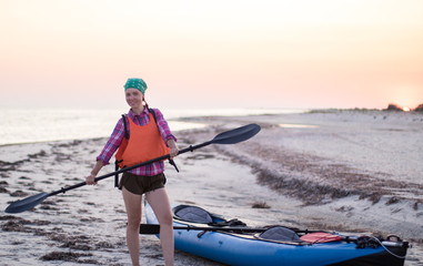 young woman with paddle near expedition kayak on the beach at sunset time