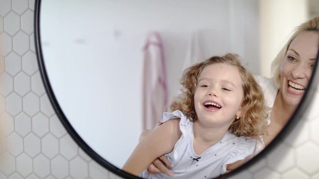 A cute small girl with mother in bathroom indoors at home, looking in mirror.