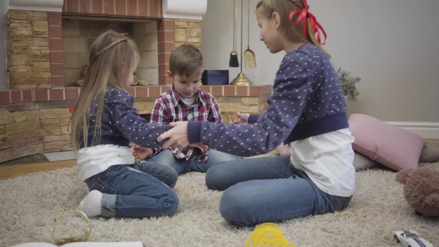 Joyful Caucasian girl teaching younger brother and sister playing a game. Happy teenager spending free time with kids at home. Unity, happiness, leisure.
