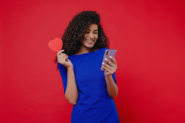 beautiful black woman with heart shaped valentine card looking at phone on red background
