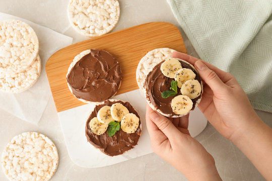 Woman holding puffed rice cake with chocolate spread and banana at grey marble table, top view