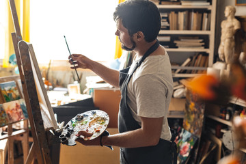 Male painter at art studio indoors with palette looking at brush inspired